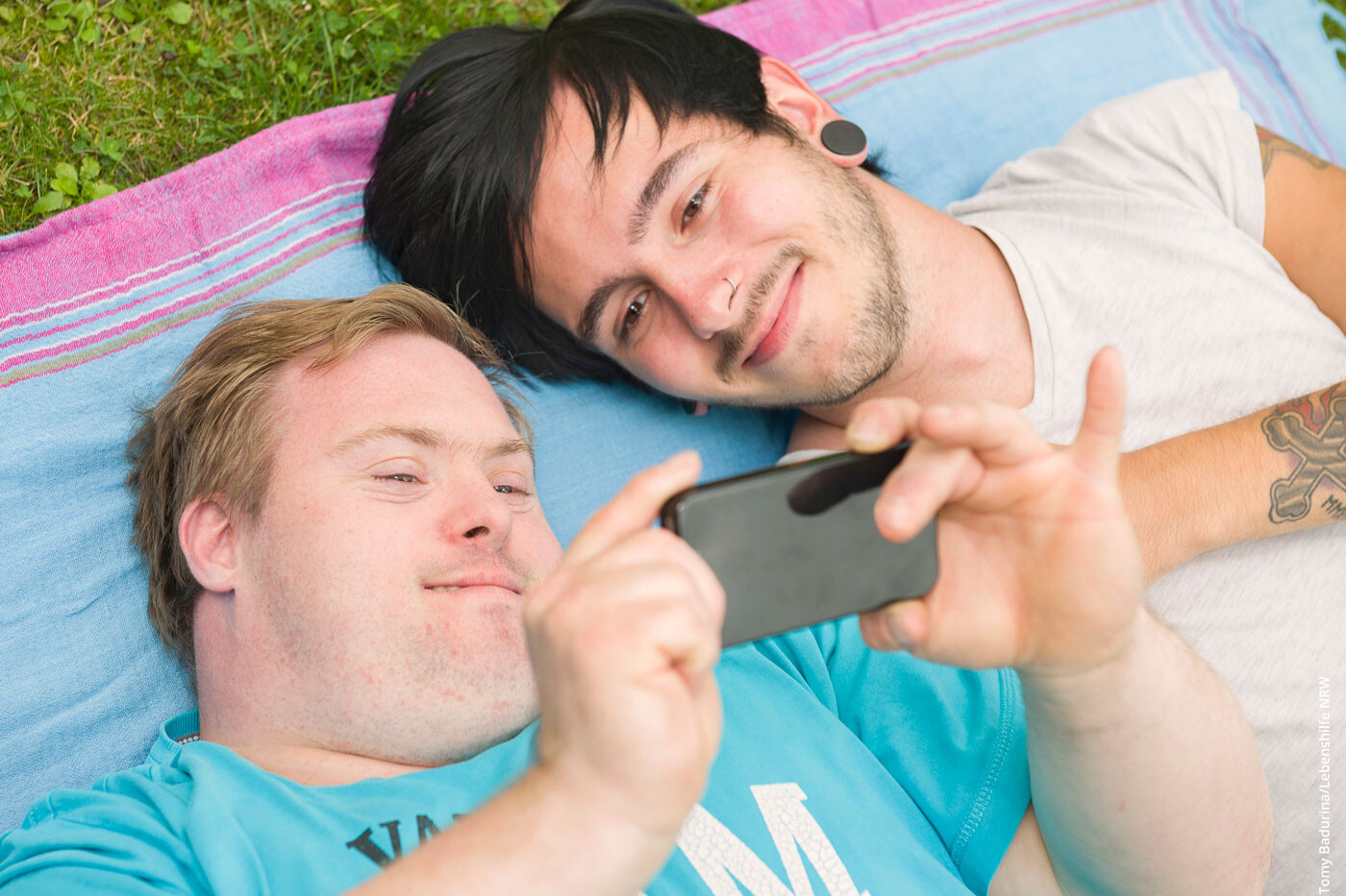 Ein junger Mann und ein junger Mann mit Down-Syndrom liegen im Gras auf einer Decke und sehen gemeinsam etwas auf einem Handy an.