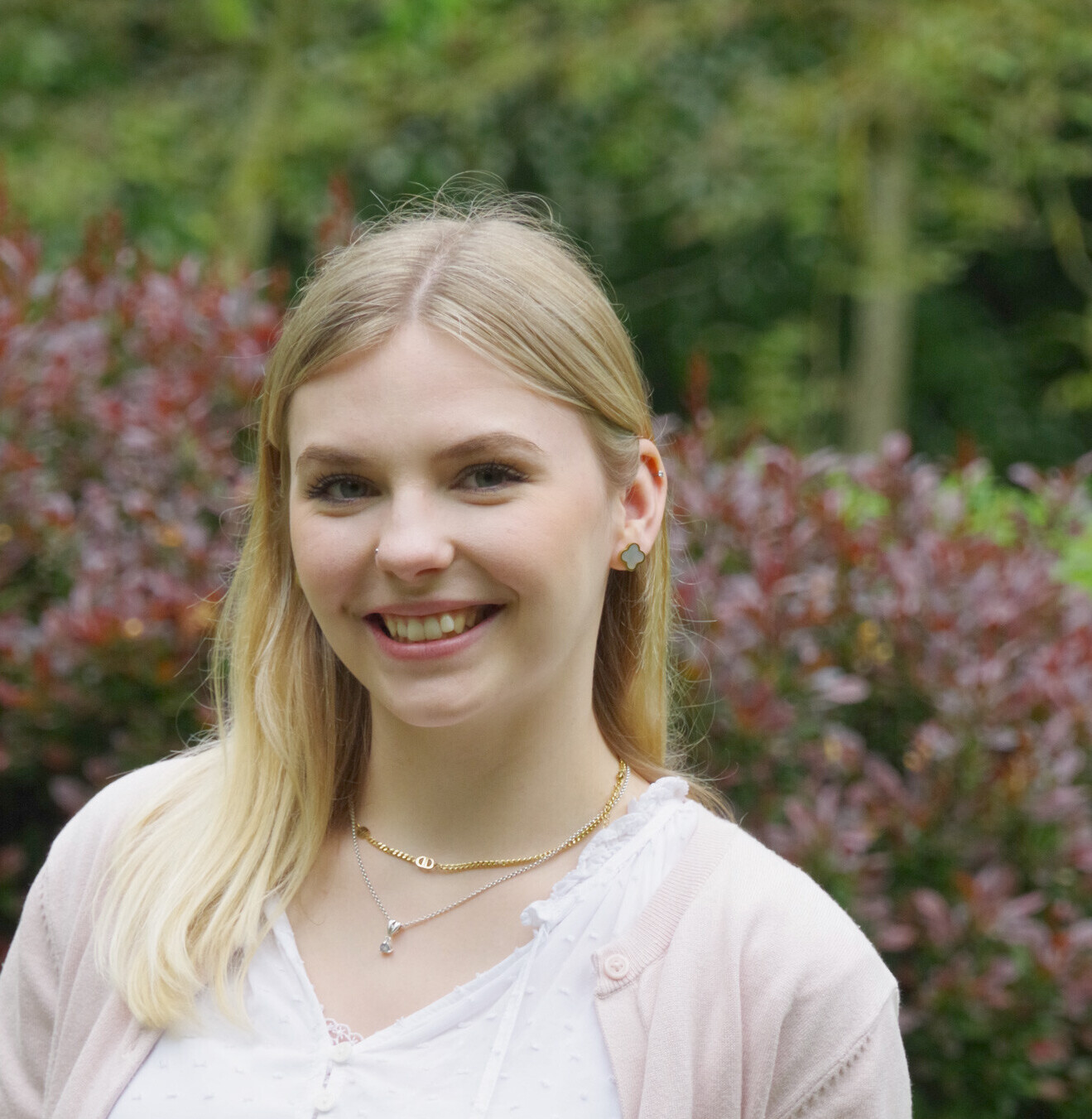 Porträt einer blonden jungen Frau vor einer Hecke und Bäumen.