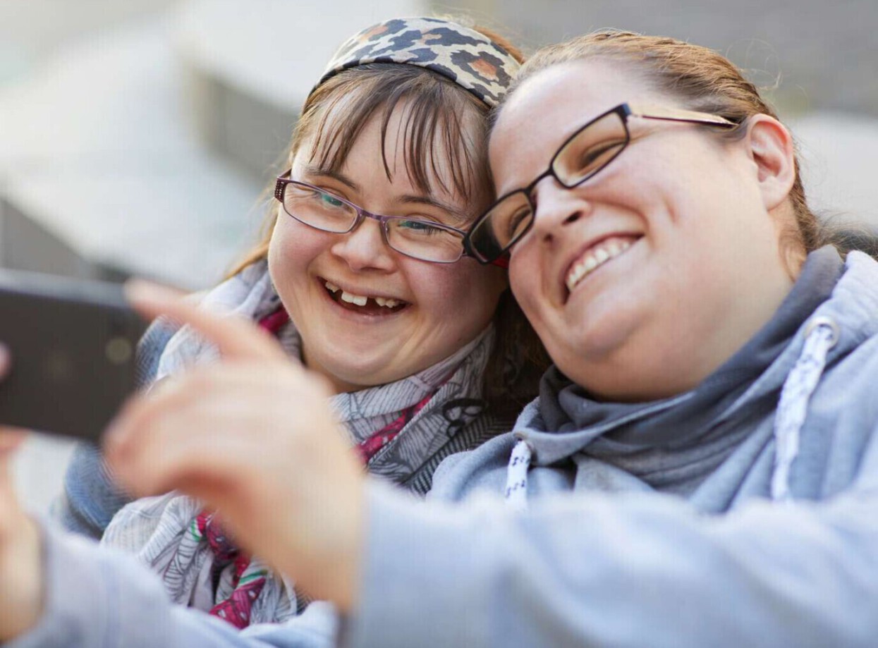 Lebenshilfe_5D4_3_F5A2783-1600px Ein Mädchen mit einer Behinderung und eine Mitarbeiterin sehen sich lachend etwas auf einem Handy an.